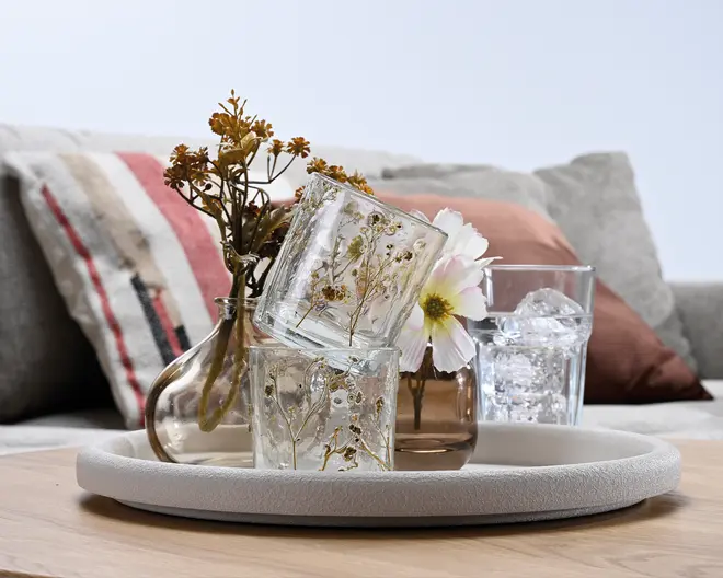 Dekorative glasvaser med pressede blomster og tørrede stilke arrangeret på et rundt fad, med et lille klart glas, en ravfarvet vase og et glas med is i baggrunden.