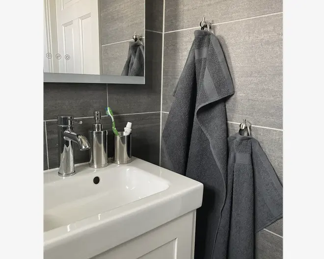 To mørkegrå håndklæder med en diskret kantdetalje hænger på kromkroge mod en grå flisevæg på badeværelset. Håndklæderne ser bløde og fyldige ud og har et moderne, minimalistisk design, der passer til det stilrene badeværelse.