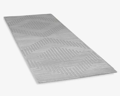 Rektangulært tæppe i lysegrå farve med et diskret, hævet geometrisk mønster af diagonale linjer, der danner chevron-former. Teksturen giver et moderne og minimalistisk udtryk. Kanterne er pænt afsluttede, og overfladen ser blød og lavluvet ud, hvilket gør det velegnet til områder med meget trafik.