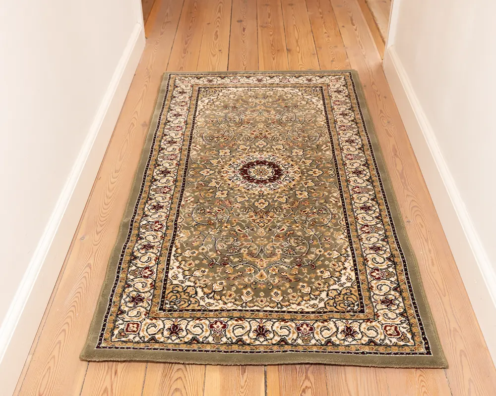 Rektangulært tæppe med traditionelt persisk-inspireret design, der har et centralt medaljonmønster omgivet af indviklede blomster- og vinrankemotiver i afdæmpede grønne, beige og bordeaux nuancer. Tæppet har en bred dekorativ kant med gentagne blomstermotiver og en let forhøjet tekstur. Det ligger på et lyst trægulv i en gang.