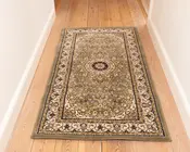Rektangulært tæppe med traditionelt persisk-inspireret design, der har et centralt medaljonmønster omgivet af indviklede blomster- og vinrankemotiver i afdæmpede grønne, beige og bordeaux nuancer. Tæppet har en bred dekorativ kant med gentagne blomstermotiver og en let forhøjet tekstur. Det ligger på et lyst trægulv i en gang.