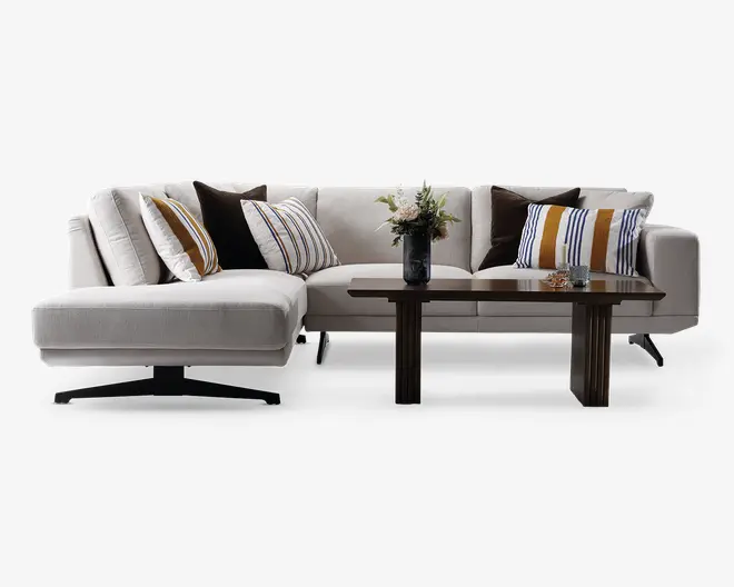 Moderne L-formet sofa med lyse beige betræk, brede sæder og bløde ryghynder. Sofaen har flere pyntepuder med stribede mønstre i hvid, sennepsgul og mørkeblå samt ensfarvede mørkebrune puder. Foran siddeområdet står et rektangulært sofabord i træ med en vase med blomster og små dekorative genstande.