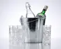 En blank metal isspand indeholder to flasker, én klar med svinglåg og én grøn med korkprop og etiket. Rundt om spanden står fire høje, klare glas, alle fyldt med isterninger. Glassene har et facetslebet, mønstret design nær bunden, hvilket giver et dekorativt udtryk.
