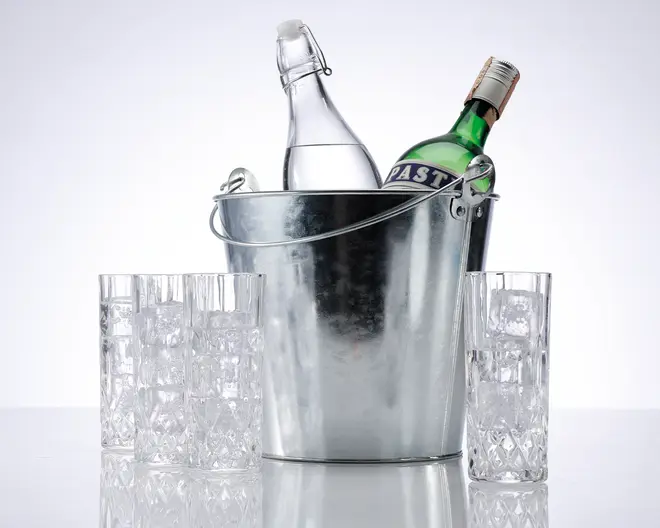 En blank metal isspand indeholder to flasker, én klar med svinglåg og én grøn med korkprop og etiket. Rundt om spanden står fire høje, klare glas, alle fyldt med isterninger. Glassene har et facetslebet, mønstret design nær bunden, hvilket giver et dekorativt udtryk.