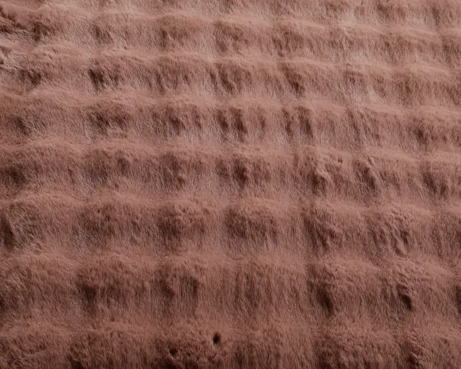 Blødt, plysset stof med en tekstureret, gitterlignende mønster. Materialet virker tykt og hyggeligt med hævede riller og fordybninger, der skaber en quiltet effekt. Farven er en varm, støvet rosa eller lysebrun, og overfladen ser indbydende og behagelig ud, ideel til brug i sengetøj eller tæpper.