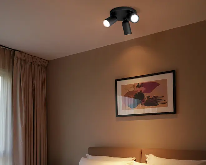 Sort loftspot med tre justerbare hoveder, hvor to lys er tændt, monteret i et beige soveværelsesloft over et indrammet abstrakt billede og en seng.