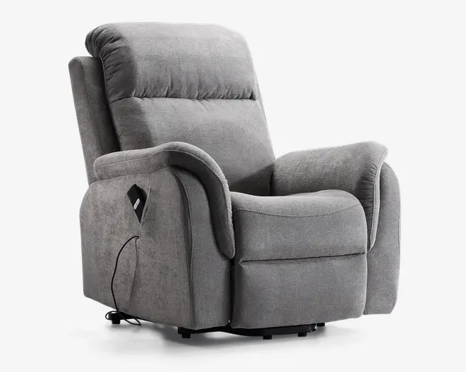 Blød, grå polstret lænestol med tykt polstret ryglæn, sæde og armlæn for ekstra komfort. Har en sidelomme til opbevaring af fjernbetjening samt en tilsluttet ledning, hvilket indikerer elektrisk recliner- eller massagefunktion. Stolen har et moderne, indbydende design med blødt stof og let buede former.