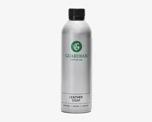 En høj, cylindrisk sølvfarvet flaske med sort skruelåg. Etiket med grønt, cirkulært logo med et 'G' og teksten 'GUARDIAN Furniture Care' i grøn skrift. Under logoet står der 'LEATHER SOAP' med store sorte bogstaver, og nederst er der oversættelser til flere sprog. Flasken har et stilrent, moderne design og ser ud til at være lavet af metal eller metallisk plast.
