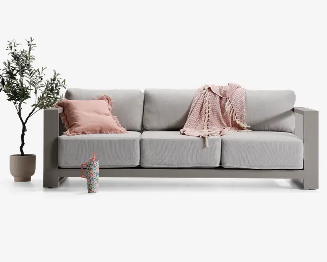 En moderne tresæders sofa med lysegrå, tykke sæde- og ryghynder. Sofaen har brede, firkantede armlæn og en solid ramme. Den er pyntet med et blødt, lyserødt tæppe med frynser, der er lagt over den ene side, samt en matchende lyserød pyntepude med rynkede kanter. Ved siden af sofaen står en potteplante med grønne blade, og foran sofaen står en farverig, mønstret drikkekop med sugerør.