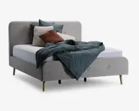 En moderne seng med polstret stel i lysegråt stof, polstret hovedgærde og fodgærde med knapdetaljer. Sengen står på fire skrå træben i naturfarve. Sengen er redt med en hvid madras, et mørkeblåt dynebetræk, der hænger ud over kanten, samt flere pyntepuder i nuancer af blågrøn, brun og creme, herunder en tekstureret cremefarvet pude for ekstra kontrast.