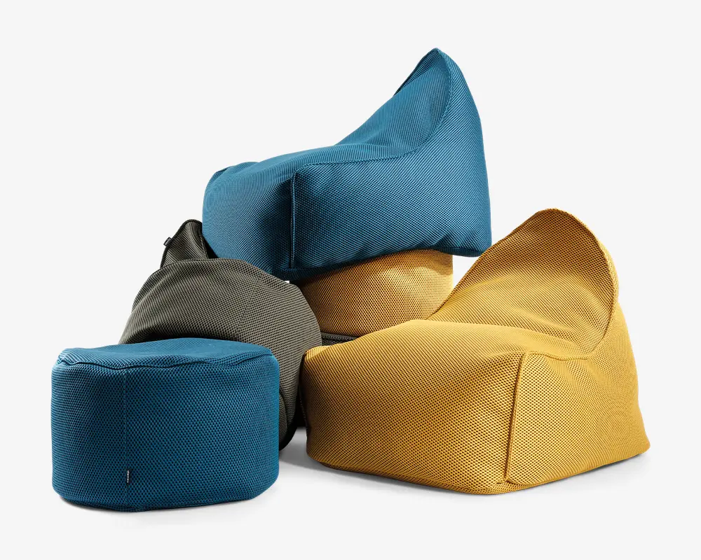 En gruppe på fire bløde, stofbetrukne siddepladser i forskellige former og farver. Sæderne inkluderer en blå kubeformet puf, en gul loungestol med skrånende ryglæn, en blå stol med høj ryg og et gråt bønneformet sæde. Stoffet har en tekstureret, vævet overflade, og farverne er klare, hvilket gør dem velegnede til moderne eller afslappede opholdsrum.