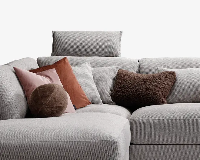 Nærbillede af en moderne, lysegrå stofsofa med forskellige pyntepuder. Puderne inkluderer en rund, blød brun pude, en firkantet brun pude med tekstur, en rustfarvet pude og to bløde, lyserøde puder, alle i forskellige former og materialer. Betrækket har en diskret vævet struktur, og sædet ser dybt og komfortabelt ud med en støttende ryg.