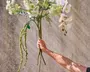 En hånd holder en buket kunstige blomster med lange stilke. Arrangementet består af en blanding af hvide blomster, herunder orkidéer og andre fine blomster, samt grønt løv og hængende grønne elementer. Blomsterne har et realistisk udseende med detaljerede kronblade og blade. Baggrunden er en enkel, tekstureret væg, som fremhæver blomsterarrangementets detaljer og naturtro kvalitet.