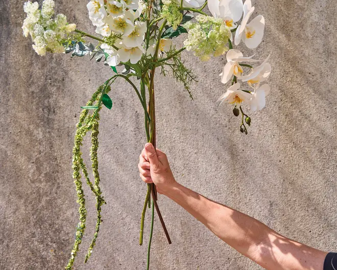 En hånd holder en buket kunstige blomster med lange stilke. Arrangementet består af en blanding af hvide blomster, herunder orkidéer og andre fine blomster, samt grønt løv og hængende grønne elementer. Blomsterne har et realistisk udseende med detaljerede kronblade og blade. Baggrunden er en enkel, tekstureret væg, som fremhæver blomsterarrangementets detaljer og naturtro kvalitet.