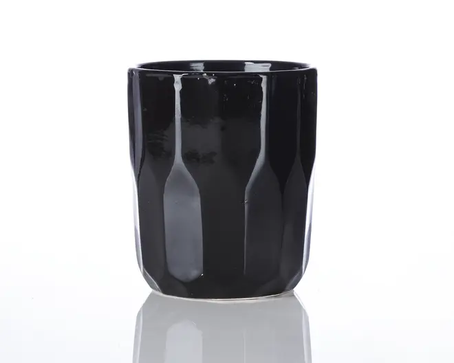 En blank sort kop med en facetteret, geometrisk overflade. Koppen har en cylindrisk form med let tilspidsede sider og en glat kant. Ydersiden har lodrette riller, der giver tekstur og visuel interesse. Bunden er flad, og det samlede design er moderne og minimalistisk.