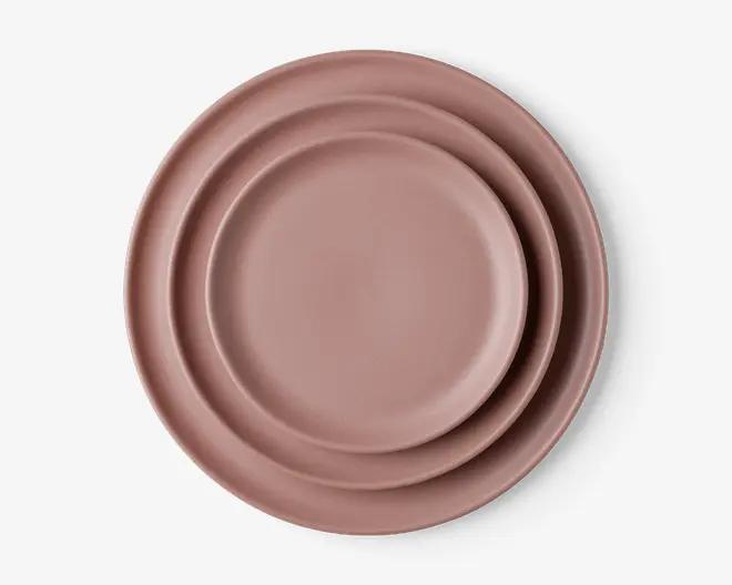 Sæt med tre runde tallerkener i forskellige størrelser, alle i en mat soppefarvet rosa. Tallerkenerne har et enkelt, minimalistisk design med glat overflade og let hævede kanter. De er stablet pænt fra den mindste til den største |.