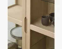 Nærbillede af et vitrineskab i lyst træ med glaslåger og glashylder. Skabet har et lodret indfræset greb, der gør det nemt at åbne. Indeni står keramikkopper og tallerkener pænt arrangeret, hvilket fremhæver det rummelige og organiserede interiør. Træets åretegning er synlig og giver et naturligt og moderne udtryk.