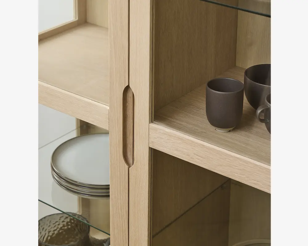 Nærbillede af et vitrineskab i lyst træ med glaslåger og glashylder. Skabet har et lodret indfræset greb, der gør det nemt at åbne. Indeni står keramikkopper og tallerkener pænt arrangeret, hvilket fremhæver det rummelige og organiserede interiør. Træets åretegning er synlig og giver et naturligt og moderne udtryk.
