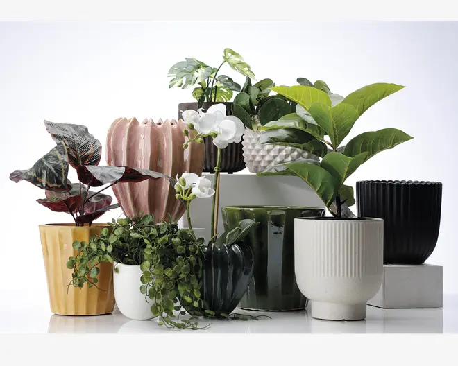 Et udvalg af dekorative urtepotter i forskellige former, størrelser og farver er udstillet sammen. Potterne har teksturerede overflader, herunder riflede, rillede og facetterede designs, og findes i nuancer af gul, hvid, lyserød, grøn, sort og mørkebrun. Nogle potter har en blank finish, mens andre er matte. Flere potter indeholder kunstige planter med frodige grønne blade, hængende ranker og hvide orkidéblomster, hvilket giver arrangementet et livligt og naturligt udseende.