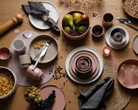 Et sæt keramikservice i afdæmpede rosa, beige og cremefarver, herunder tallerkener, skåle og kopper. Delene har en mat overflade og et minimalistisk design. Nogle skåle er fyldt med frisk frugt, pasta eller druer, mens andre er stablet eller indeholder sammenrullede stofservietter. Arrangementet indeholder bestik i rustfrit stål, en peberkværn og dekorative elementer som tørrede blomster og kaffebønner spredt på et træbord. Stilen er moderne og elegant, velegnet til både afslappet og formel servering.