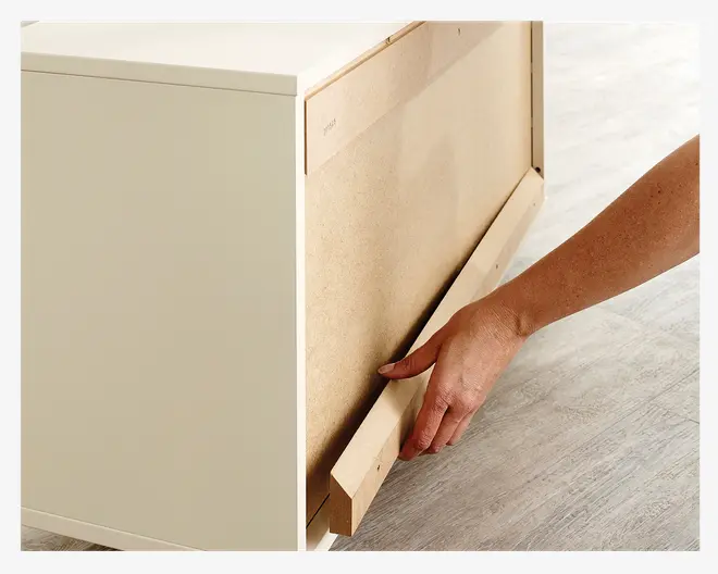 Nærbillede af en person, der samler et møbel, hvor en hånd fastgør eller justerer en træstøttelist på bagpanelet. Møblet har en lys overflade og står på et lyst trægulv. Billedet fremhæver samlingsprocessen og detaljerne i bagpanelets og støttelistens konstruktion.