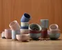 En samling keramiske kopper og skåle i forskellige pastelfarver, herunder rosa, blå, grå og creme. Produkterne har en mat overflade og et minimalistisk design, hvor nogle har enten ensfarvede eller let prikkede mønstre. Sættet består af både cylindriske og runde former, og flere af delene er stablet eller vippet for et afslappet, kunstnerisk udtryk. Nogle skåle indeholder kaffebønner, og en tepose ses ved siden af en af kopperne, hvilket understreger deres anvendelse til varme drikke. Det samlede udtryk er moderne og stilfuldt, velegnet til et nutidigt borddækning.
