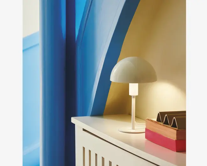 En lille bordlampe med et minimalistisk design, der har en kuppelformet mat hvid skærm og en slank cylindrisk fod. Lampen står på en lys overflade ved siden af en brevholder og en rød bog. Lampen udsender et blødt, varmt lys, der skaber en hyggelig stemning.