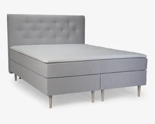 Lys grå polstret seng med høj, knap-tuftet sengegavl. Sengen har et minimalistisk design med rene linjer og står på seks koniske træben. Madrassen ligger plant med rammen, og det samlede udtryk er moderne og elegant.