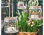 Klar glasbeholdere dekoreret med pressede blomster og botaniske motiver, hver ophængt i rustik snor. Beholderne indeholder hvide fyrfadslys, hvoraf det ene er tændt. Blomsterdekorationerne har gule, hvide og lilla blomster, hvilket giver et naturligt, håndlavet udtryk. Glassene hænger udendørs og egner sig som dekorative lanterner til have eller terrasse.