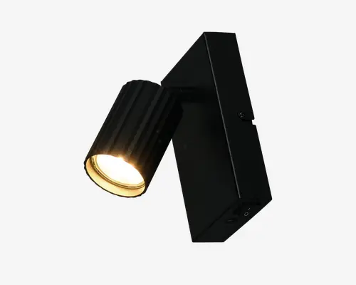 Justerbar vægmonteret spotlampe med cylinderformet, rillet lampehoved i sort metal og rektangulær sort base. Spotlampen udsender et varmt hvidt lys og kan rettes i den ønskede retning, hvilket gør den velegnet til accent- eller arbejdsbelysning. Designet er moderne og minimalistisk med en mat overflade.