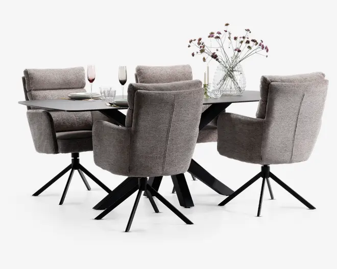 Billedet viser et moderne spisebordssæt med et rektangulært bord med mørk, mat overflade og skrå sorte metalben. Rundt om bordet står fire polstrede stole i blødt, neutralt gråt stof, hver med høj ryg, polstret sæde og armlæn for komfort. Stolene har sorte metalstel med drejefunktion og fire ben. Bordet er dækket med glas, tallerkener og en dekoration bestående af en klar glasvase med fine lilla og hvide blomster.