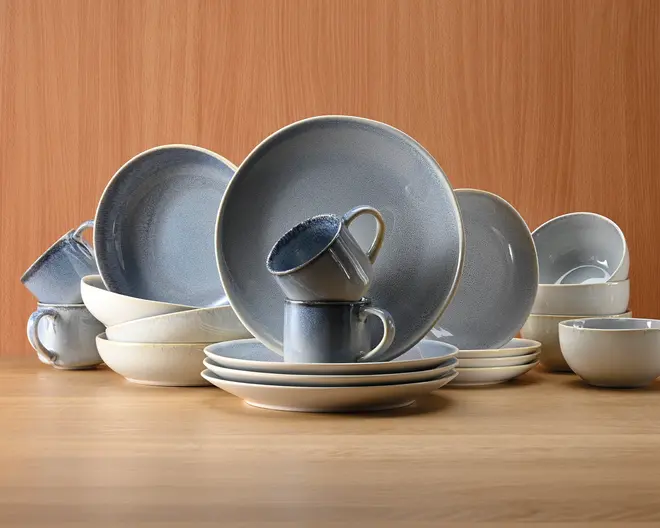 Et sæt stentøj med tallerkener, skåle og krus i et ensartet design med mat finish og farvegradient fra blød blågrå til råhvid. Sættet består af store middagstallerkener, mindre sidetallerkener, skåle og krus, alle med en diskret plettet tekstur og blødt afrundede kanter. Delene er stablet og arrangeret, så deres ensartede farve og håndlavede udtryk fremhæves, velegnet til både hverdags- og festbrug.