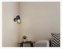 En moderne vægmonteret lampe med en cylindrisk sort metalskærm, der har et netdesign, som giver et blødt lys. Lampen er fastgjort til en rund sort base og er placeret over et lille sidebord. Bordet har en lille potteplante (sukkulent) og et par bøger. Indretningen inkluderer en lysefarvet sofa med en sort og hvid chevron-mønstret pude, hvilket skaber en moderne og hyggelig atmosfære.