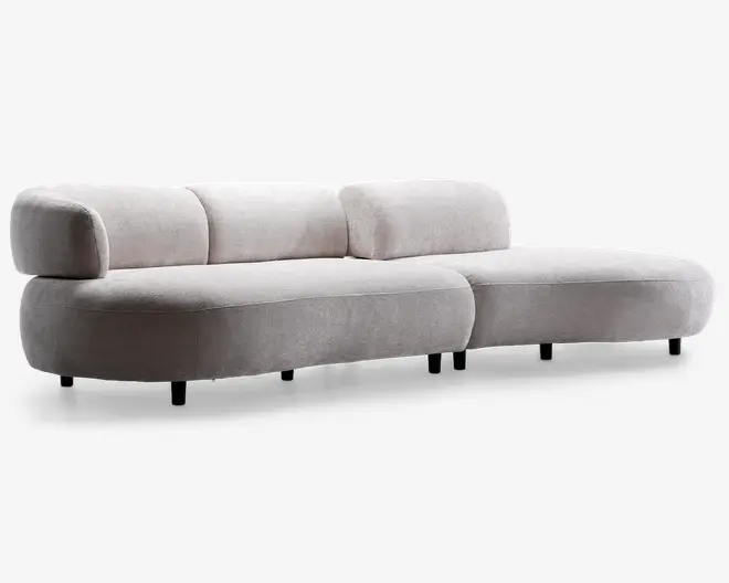 Moderne sofa med lav profil, afrundede kanter og bløde, fyldige hynder både i sæde og ryg. Betrukket med et lyst beige stof med let struktur. Sofaen har et bredt siddeareal og står på lave, næsten usynlige ben, hvilket giver et svævende udtryk. Udtrykket er minimalistisk og nutidigt, velegnet til moderne indretning.