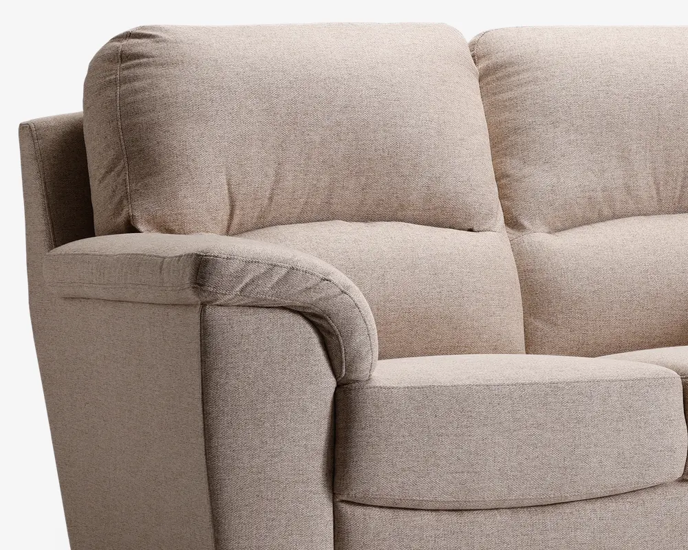 Nærbillede af et sofahjørne med bløde, fyldige ryg- og sædehynder for ekstra komfort. Betrækket er i en lys beige tekstil med diskret struktur. Armlænet er afrundet og polstret, hvilket giver ekstra støtte. Syningerne er synlige langs kanterne, hvilket vidner om solid konstruktion.