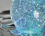 Nærbillede af en kugleformet genstand fyldt med glitrende blå og sølvfarvet glimmer, der skaber en skinnende effekt. Overfladen er glat og blank, og genstanden hviler på en reflekterende, metallisk base med kronbladslignende former. Glimmeret indeni kuglen reflekterer lyset og giver et levende, iriserende udseende.