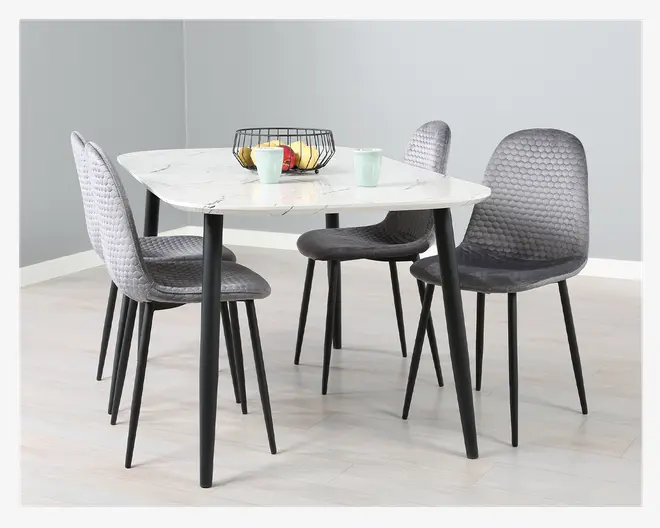 Et moderne spisesæt med et rektangulært bord med hvid marmoreffekt bordplade og sorte skrå ben. Sættet inkluderer fire stole med polstrede sæder og rygge, betrukket med gråt stof med bikubemønster. Stolenes ben er sorte og let tilspidsede. Bordet er dekoreret med en trådkurv med æbler og bananer samt to pastelfarvede krus.