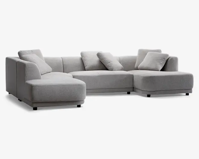 En moderne, L-formet sofa med en venstrevendt chaiselong. Sofaen er betrukket med lysegråt stof og har et stilrent design med lave armlæn og ryglæn. Der medfølger fem matchende, firkantede puder for ekstra komfort. Sædehynderne er tykke og bløde, hvilket giver god siddeplads til flere personer.