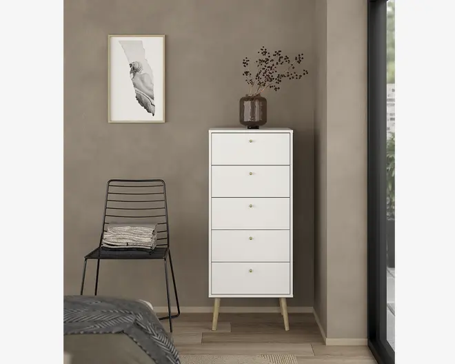 Høj, smal kommode med fem skuffer, udført i mat hvid. Hver skuffe har en lille rund gylden knop for nem åbning. Kommoden står på fire skråtstillede, lyse træben, hvilket giver et moderne, skandinavisk udtryk. Øverst står en mørk glasvase med minimalistiske tørrede grene.