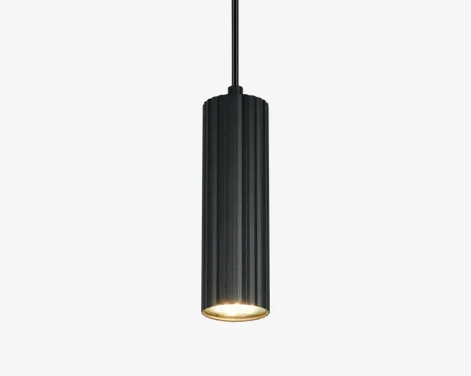 En moderne, cylindrisk pendellampe med lodrette riller i sort metal. Lampen har et slankt, aflangt design og sender lyset direkte nedad for at skabe fokuseret belysning. Den indvendige overflade nær pæren er lysere, hvilket forstærker lysets styrke. Pendlen hænger i en tynd sort ledning.