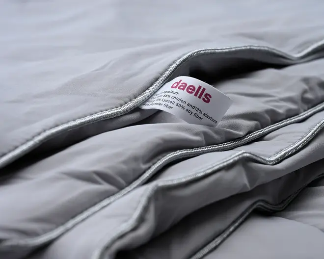 Nærbillede af et lysegråt tekstil med synlig søm og vaskeanvisning. Stoffet ser blødt og glat ud med pænt syede kanter. Mærket viser brandnavn og materialekomposition, hvilket indikerer en blanding af materialer for komfort og holdbarhed. Produktet fremstår som et velkonstrueret og kvalitetspræget tekstil.