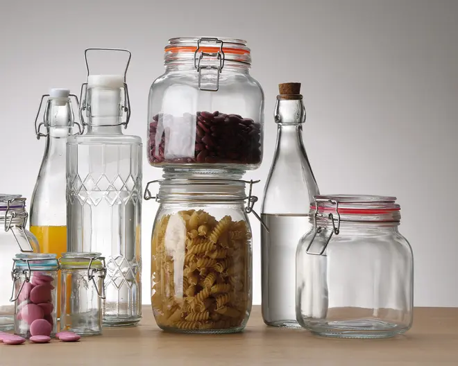 Et udvalg af klare glasopbevaringskrukker og flasker med tætsluttende låg vises. Nogle krukker indeholder tørvarer som pastaskruer, røde kidneybønner og lyserøde chokoladeknapper, mens andre er tomme. Flaskerne har patentpropper, og en indeholder en gul væske, muligvis juice, mens en anden har en korkprop. Krukkerne har orange eller blå gummipakninger for lufttæt lukning, og glasset er tykt og robust, velegnet til opbevaring i spisekammeret.