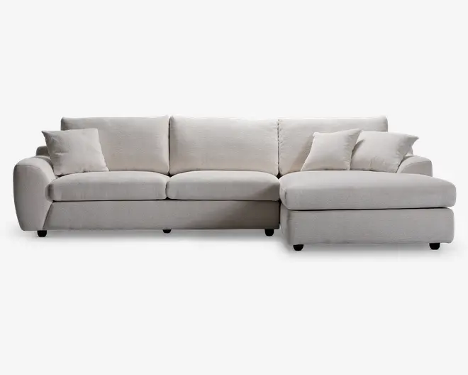 En moderne, L-formet sofa med chaiselong på højre side. Betrækket er i et lyst beige stof med en blød, tekstureret overflade. Sofaen har tre store sædehynder, tre matchende ryghynder og tre pyntepuder for ekstra komfort. Armlænene er brede og let afrundede, hvilket giver møblet et moderne og indbydende udtryk.