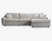 En moderne, L-formet sofa med chaiselong på højre side. Betrækket er i et lyst beige stof med en blød, tekstureret overflade. Sofaen har tre store sædehynder, tre matchende ryghynder og tre pyntepuder for ekstra komfort. Armlænene er brede og let afrundede, hvilket giver møblet et moderne og indbydende udtryk.