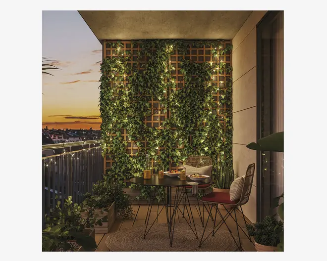 En hyggelig altanindretning ved solnedgang med en lodret træespalier dækket af frodige, grønne klatreplanter og varme lyskæder. Området har et rundt sort metalbord med stearinlys, drikkevarer og snacks, omgivet af to stole med trådramme og sædehynder. Ekstra potteplanter og et vævet tæppe bidrager til den indbydende stemning, mens lyskæder langs altanrækværket giver et blødt, stemningsfuldt skær.