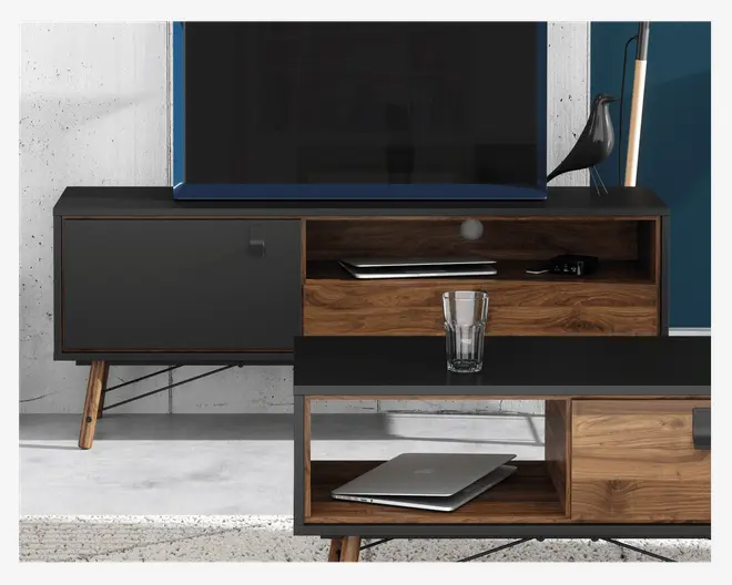 Billedet viser et moderne TV-bord og sofabord i et sæt med et retroinspireret design. Begge møbler har en kombination af mat sort og naturlig træfinish. TV-bordet har skrå træben, et lukket skab til venstre og åbne hylder til højre, hvor der står en dekorativ fuglefigur og en lampe ovenpå. Sofabordet matcher i design og har åbne hylder under bordpladen, hvor en lukket bærbar computer er placeret. Et klart glas står ovenpå sofabordet. Udtrykket er stilrent og moderne med rene linjer og praktiske opbevaringsmuligheder.