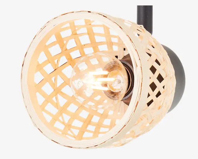 Nærbillede af en lampe med en rund, åben fletskærm i naturfarvet rattan eller flet. Skærmen har et gittermønster, der lader lyset skinne igennem og skaber en varm og hyggelig stemning. En klar pære er synlig indeni, og lampen er monteret på en sort base. Designet kombinerer et rustikt og moderne udtryk.