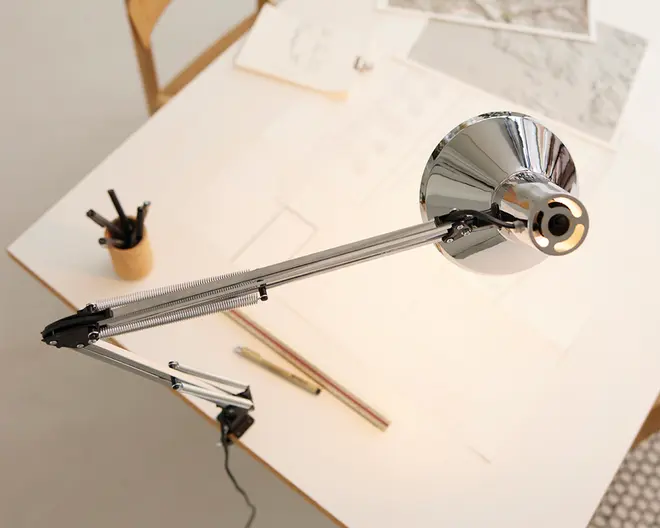 Justerbar skrivebordslampe med blank kromfinish, udstyret med en fjederbalanceret arm for fleksibel placering. Lampen er fastgjort til kanten af et skrivebord, hvilket sparer plads. Lampeskærmen er kuppelformet og reflekterende, designet til at lede lyset nedad. Armen har synlige spændingsfjedre og led, der gør det nemt at justere lampen. Lampen er placeret over en arbejdsplads med tegneredskaber, lineal og papirer, hvilket gør den velegnet til opgaver, der kræver fokuseret belysning.