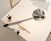 Justerbar skrivebordslampe med blank kromfinish, udstyret med en fjederbalanceret arm for fleksibel placering. Lampen er fastgjort til kanten af et skrivebord, hvilket sparer plads. Lampeskærmen er kuppelformet og reflekterende, designet til at lede lyset nedad. Armen har synlige spændingsfjedre og led, der gør det nemt at justere lampen. Lampen er placeret over en arbejdsplads med tegneredskaber, lineal og papirer, hvilket gør den velegnet til opgaver, der kræver fokuseret belysning.