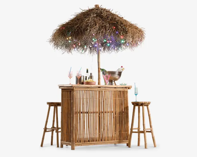 Billedet viser en bar i bambus-stil med en stråparasol over. Parasollen er pyntet med farverige lyskæder, hvilket giver en festlig stemning. Baren har to matchende bambusbarstole og er udstyret med forskellige glas, en metal isspand og cocktailtilbehør, hvilket indikerer, at den er klar til servering af drikkevarer. Det samlede udtryk er tropisk og indbydende, perfekt til udendørs sammenkomster.